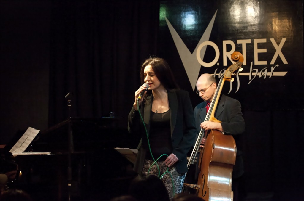 A photo of Laura Zakian and a double bass player gigging. Laura is singing in to the mic and is wearing a black blazer and patterned trousers. 