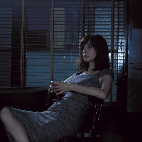 A photo of Laura sitting on a hard chair with a glass of red wine staring into the distance off camera. It is dark outside and the blinds are open.