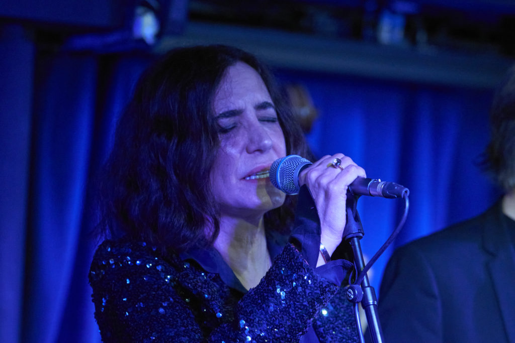 A photograph of Laura Zakian singing into the mic with her eyes closed. The background is lit in moody blues.
